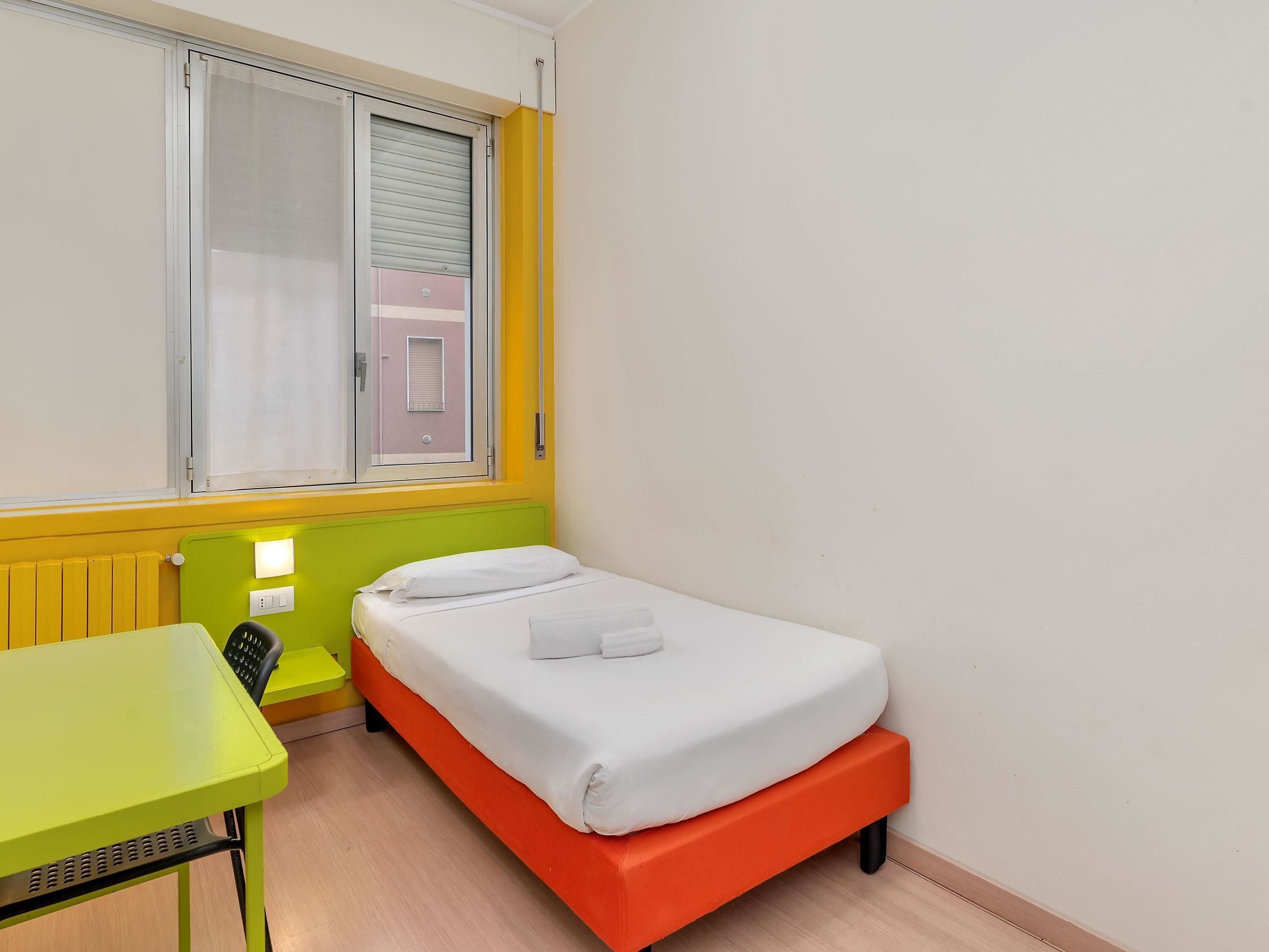 Una habitación pequeña y de colores vivos con una cama individual, una mesa verde y una ventana con persianas.