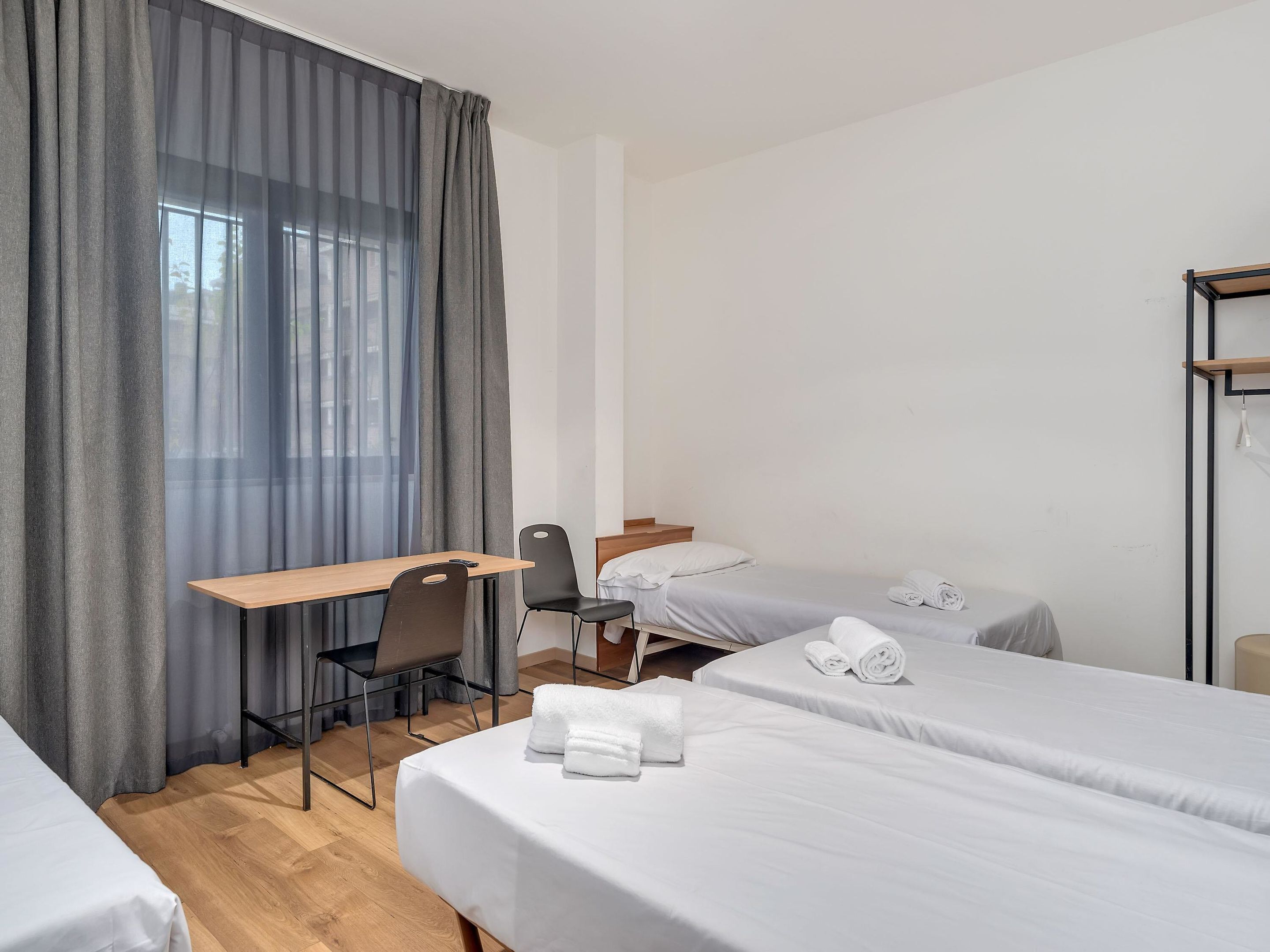 Habitación de hotel con escritorio, dos camas individuales, una silla y un amplio ventanal con cortinas.