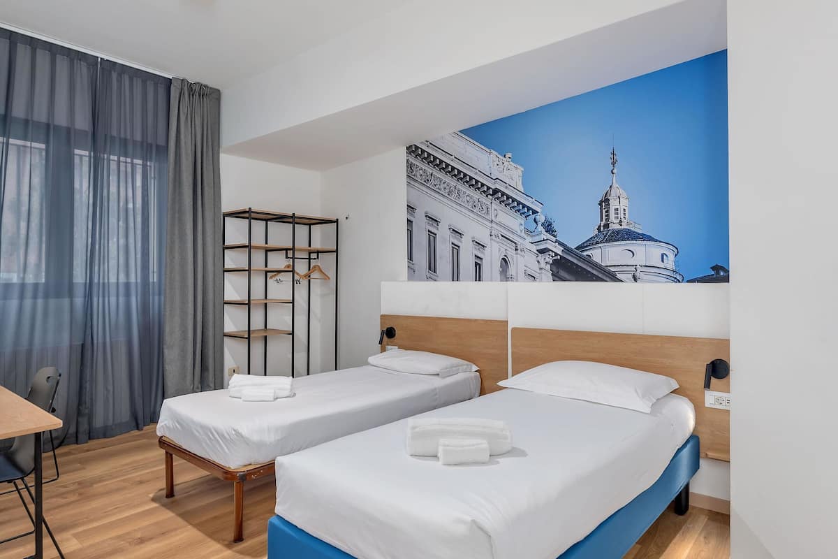 Hotellihuone, jossa on kaksi erillistä sänkyä, puinen työpöytä, tuoli, vaatekaappi ja suuri kaupunkimaisemaa esittävä seinämaalaus.