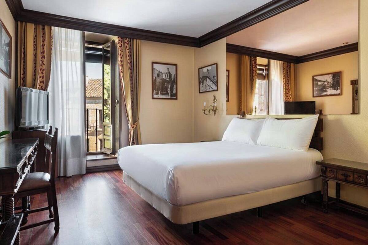 Habitación de hotel con cama, escritorio y silla. Hay cortinas, un candelabro y cuadros en las paredes.