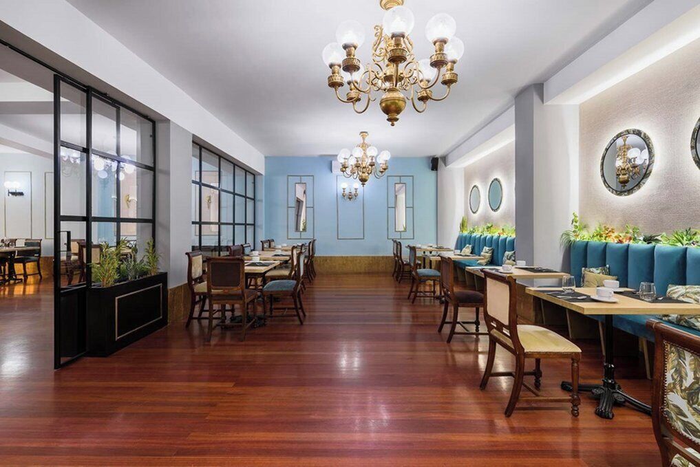 A spacious dining area with wooden flooring, multiple tables, and chairs. There are decorative chandeliers and wall-mounted mirrors.