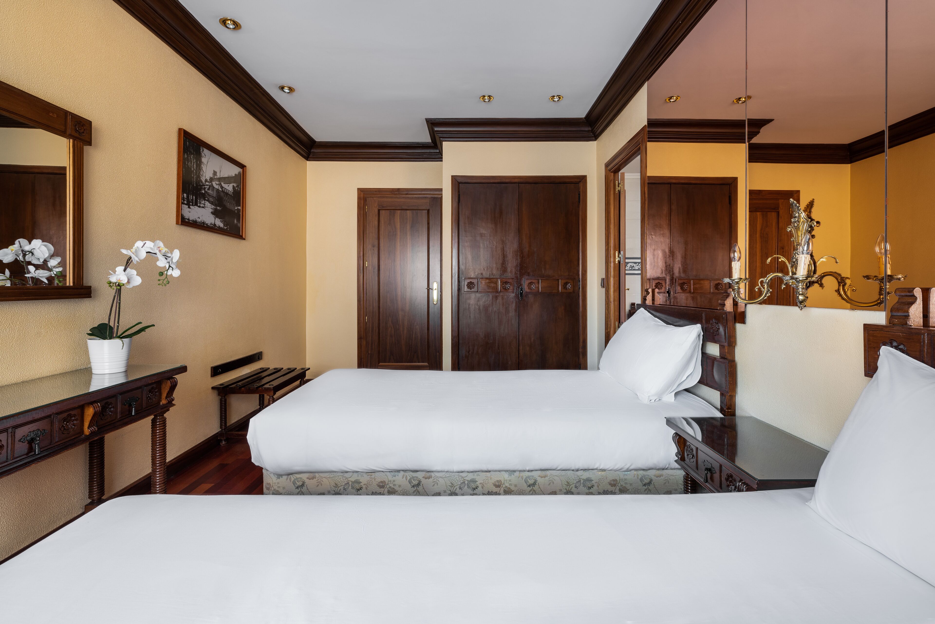 A hotel room with a large bed, a desk with a plant, a mirror, and a chandelier.