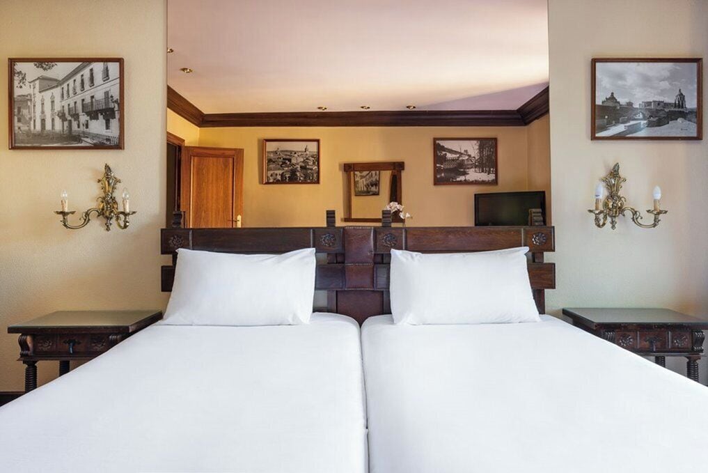 Una cama doble con ropa blanca, un cabecero de madera y una mesita de noche con una lámpara. En las paredes hay cuadros enmarcados y un televisor sobre un mueble.