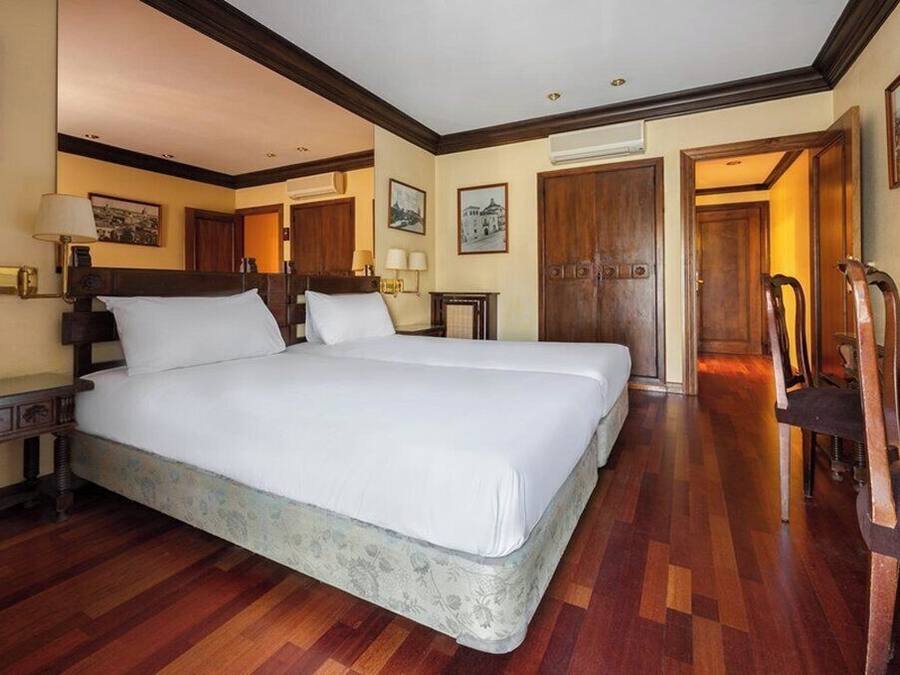 A large bed with a wooden headboard, a bedside table with a lamp, and a chair in a hotel room with wooden flooring and a wooden door.