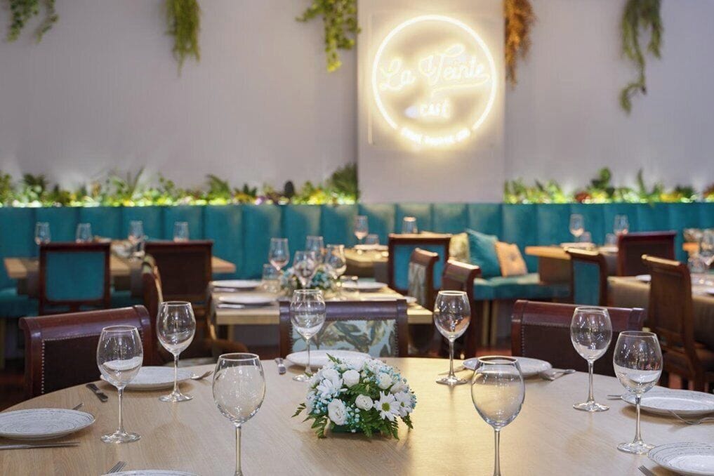 Un restaurant avec des tables dressées pour le service, un panneau néon sur le mur et des plantes vertes en arrière-plan.