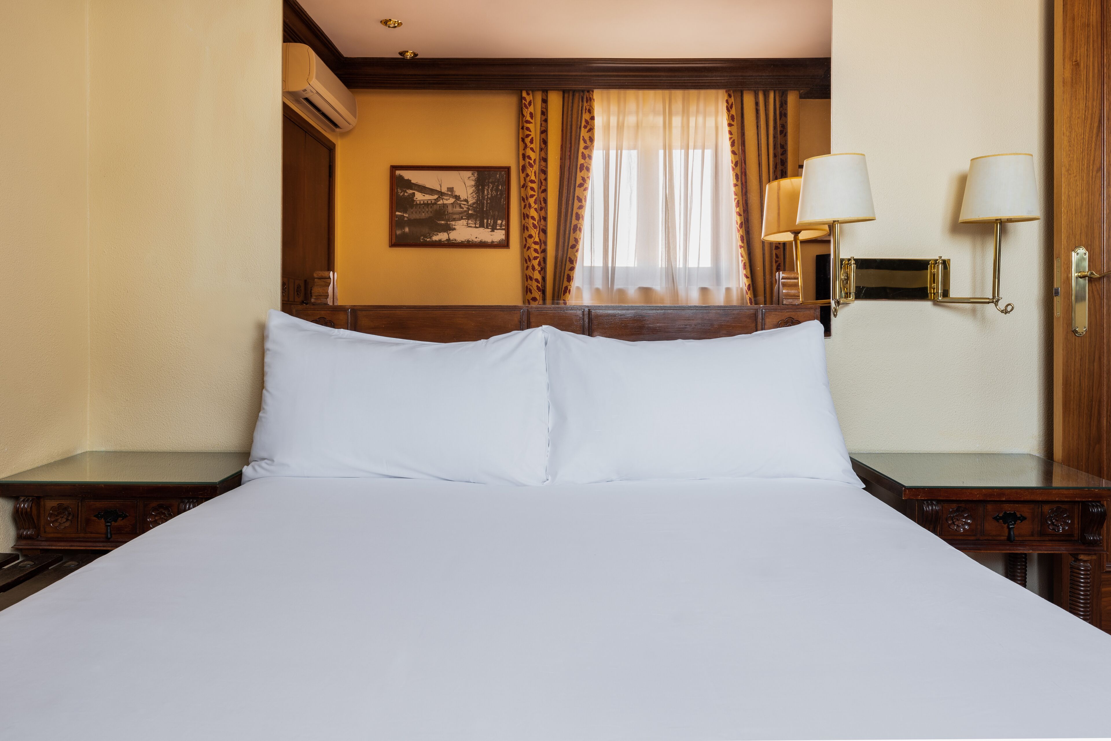 Habitació d'hotel amb un llit gran, panell de capçalera de fusta, dues taules de nit amb làmpades, una finestra amb cortines i un quadre penjat a la paret.