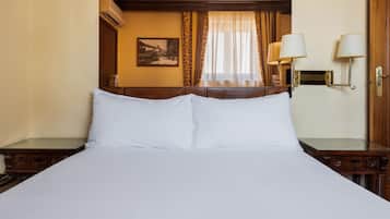 Habitació d'hotel amb un llit gran, panell de capçalera de fusta, dues taules de nit amb làmpades, una finestra amb cortines i un quadre penjat a la paret.