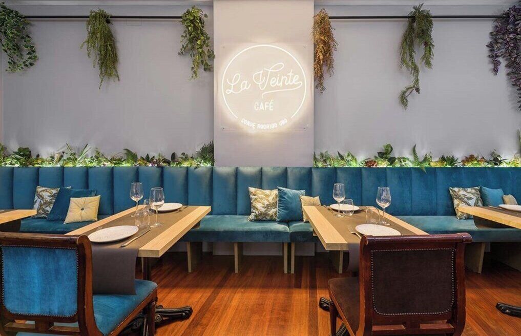 A cozy café interior with blue upholstered seating, wooden tables, and hanging plants.
