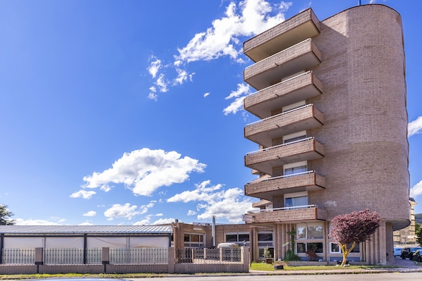 A multi-story building with a curved facade, a gated parking area, and a clear blue sky with clouds. - Hotel Vejo (Reinosa)