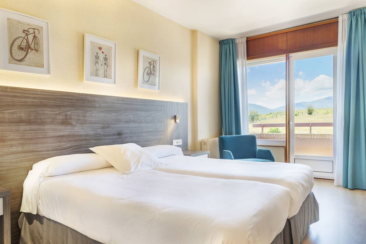 A hotel room with two beds, a view of the outdoors, and artwork on the wall.