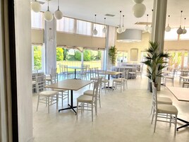 Uma ampla área de refeições interna com várias mesas e cadeiras, grandes janelas com cortinas e luminárias pendentes esféricas.