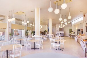 Uma sala de jantar espaçosa com várias mesas e cadeiras, tetos altos com luminárias esféricas penduradas, e uma área de cozinha visível ao fundo.