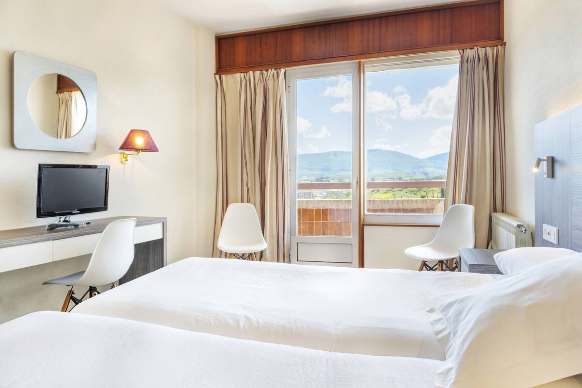 A hotel room with a bed, a desk with a computer, a chair, a TV, and a balcony with curtains.