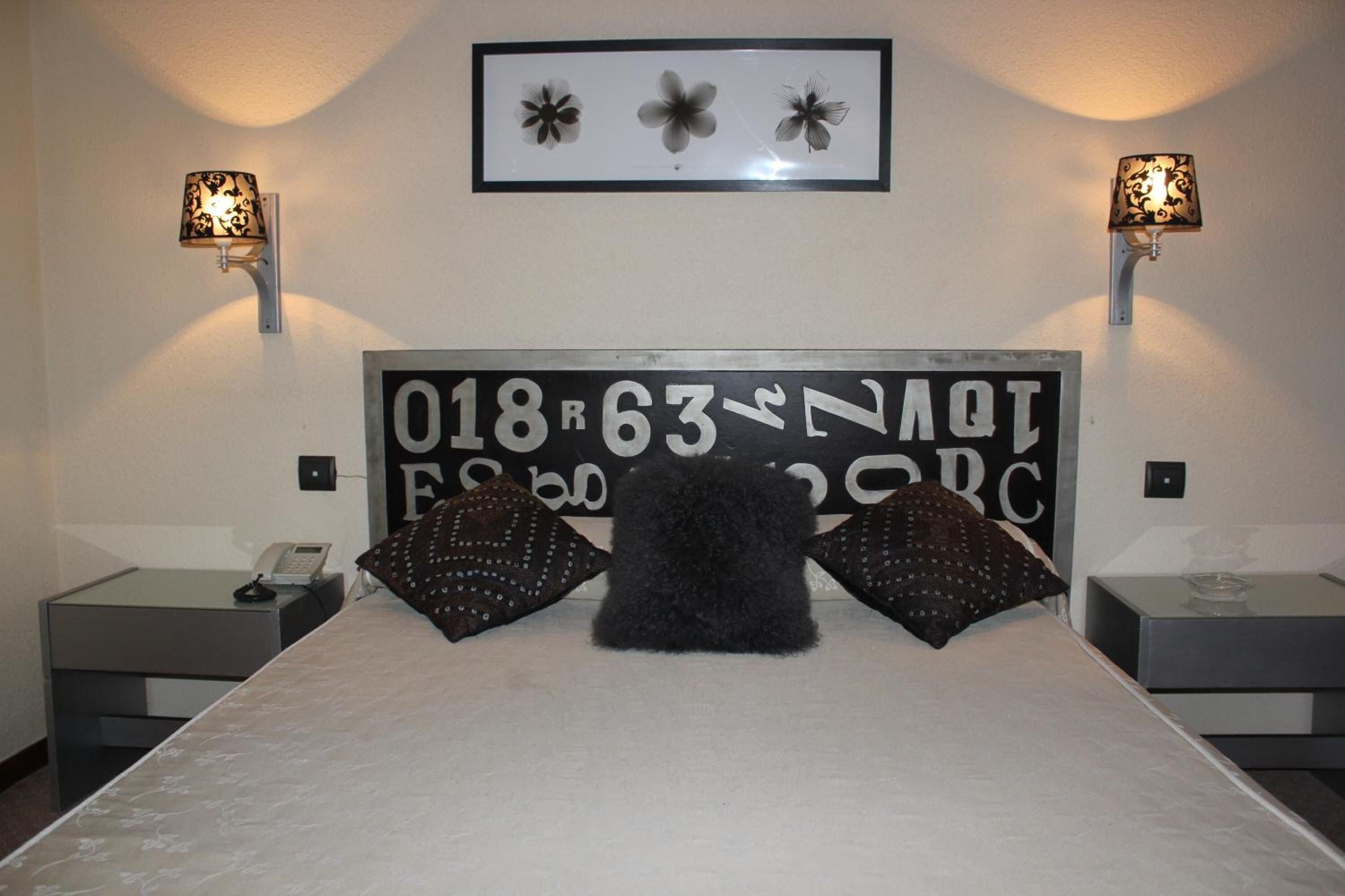 Een bed met een groot, decoratief hoofdeinde, twee wandlampen en een ingelijst schilderij erboven.