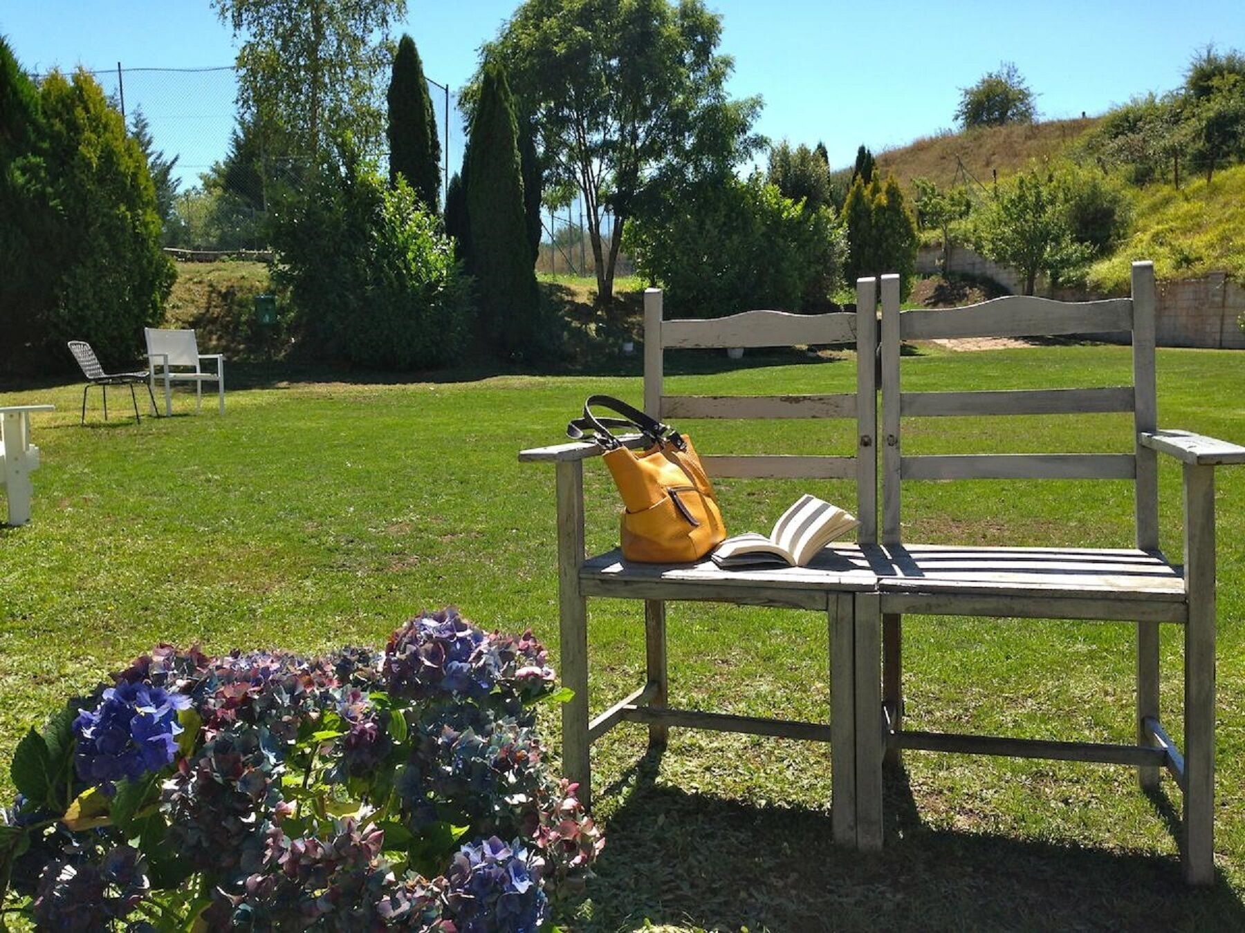A wooden bench with a yellow bag and an open book on it, surrounded by a grassy area with trees and a fence in the background.