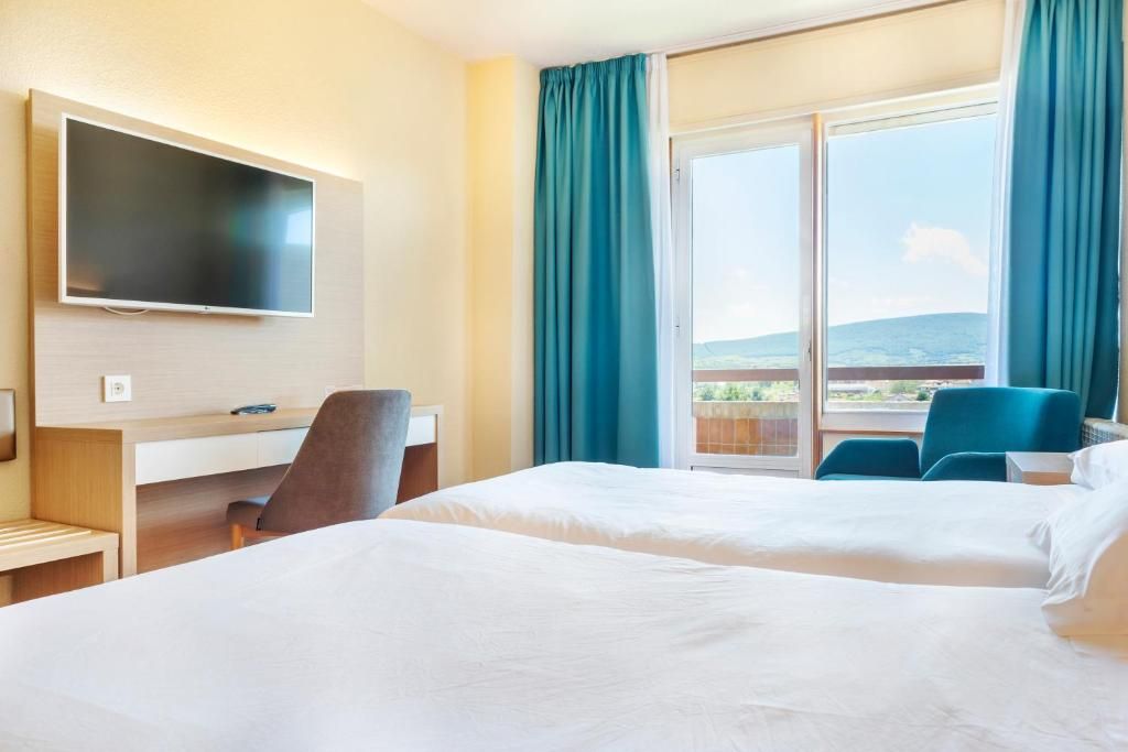 Habitación de hotel con cama, escritorio, silla, televisor y un amplio ventanal con cortinas.