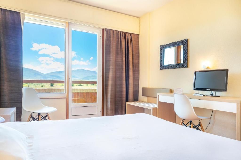 Une chambre d’hôtel comprenant un lit, deux chaises, un bureau, une télévision et une grande fenêtre avec des rideaux.