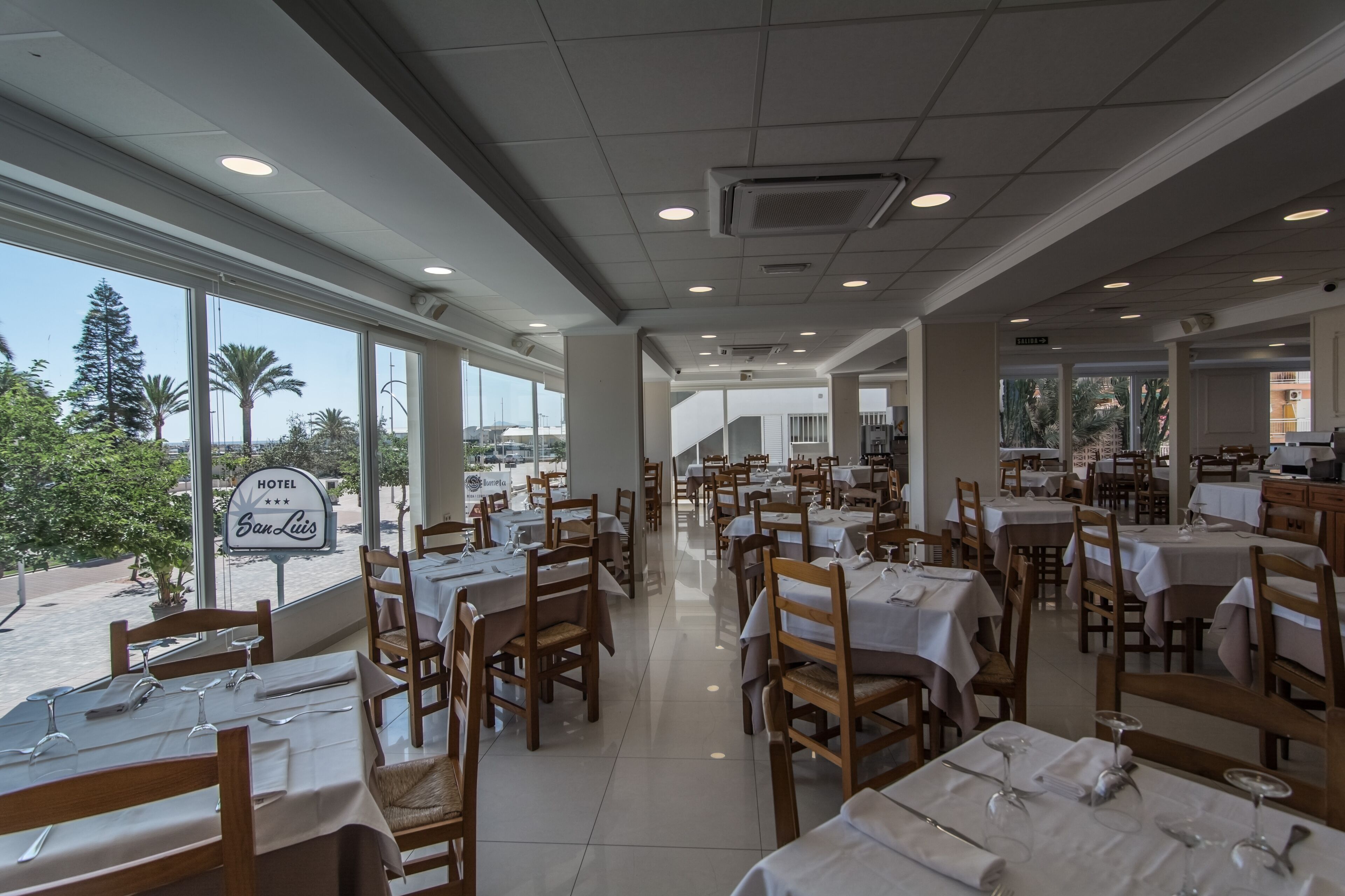 A hotel restaurant with multiple dining tables and chairs, large windows offering a view of palm trees and a clear sky.