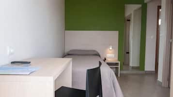 Una habitación pequeña y moderna con cama, escritorio y silla. Hay un mapa en el escritorio y una lámpara en la mesita de noche.