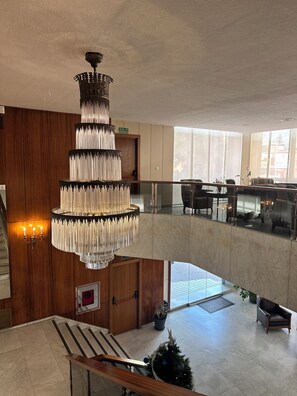A multi-level hotel lobby with a large chandelier, wooden paneling, a reception desk, and a seating area with a view of the city. - Hotel Mir Octavio (Algeciras)