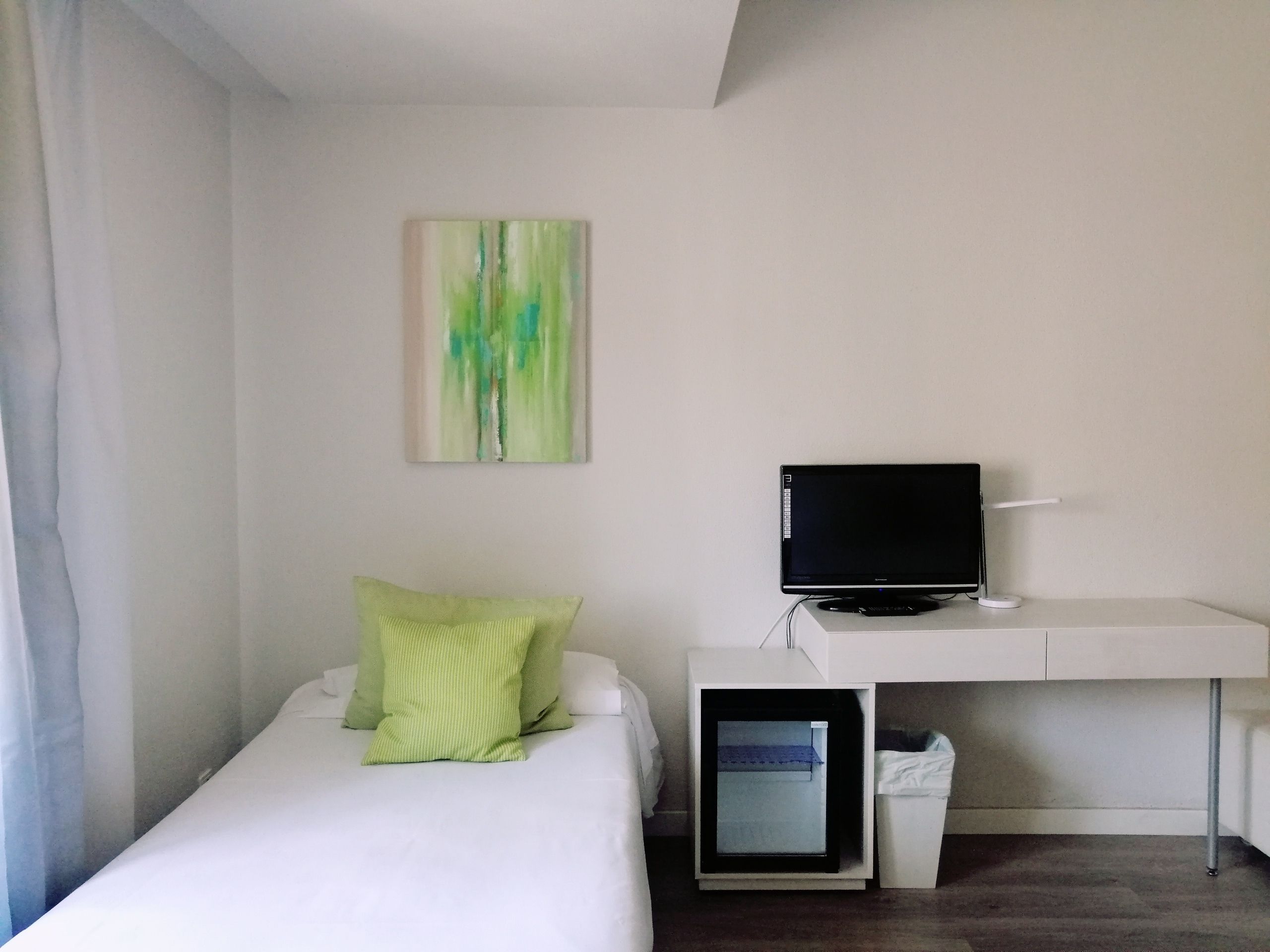 A hotel room with a bed, a desk with a computer, a trash bin, and a painting on the wall.