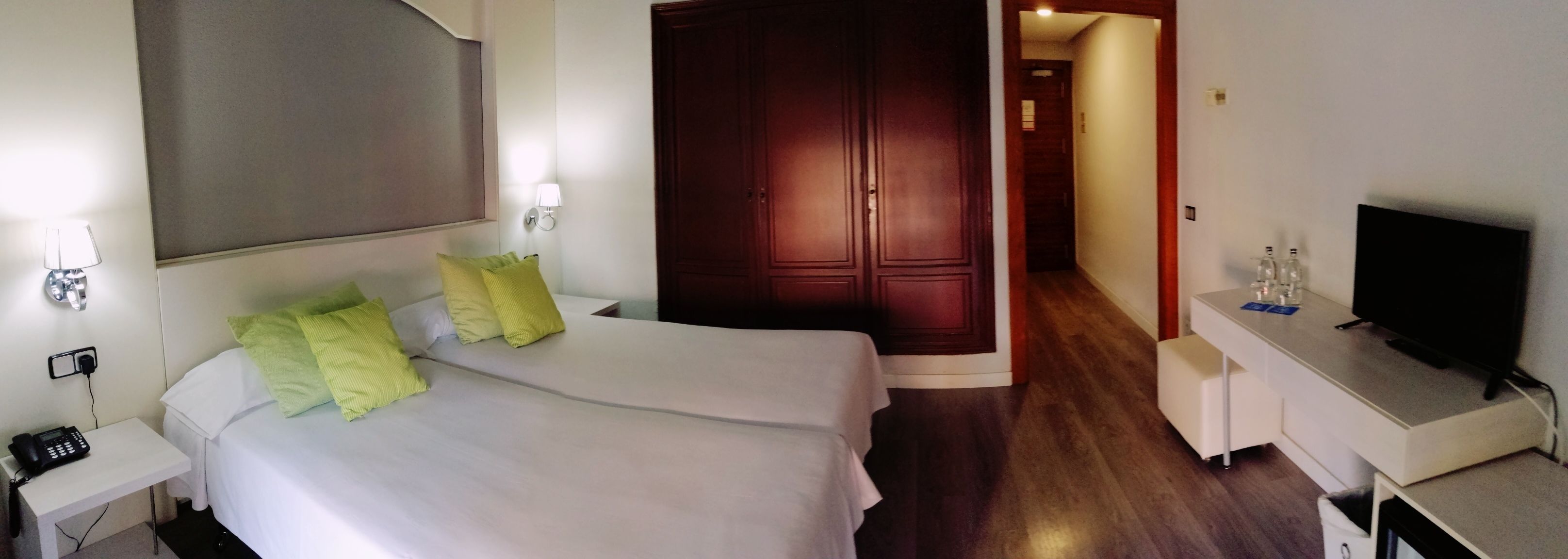 Quarto de hotel com cama, mesa de cabeceira com telefone, escrivaninha com televisão e guarda-roupa.