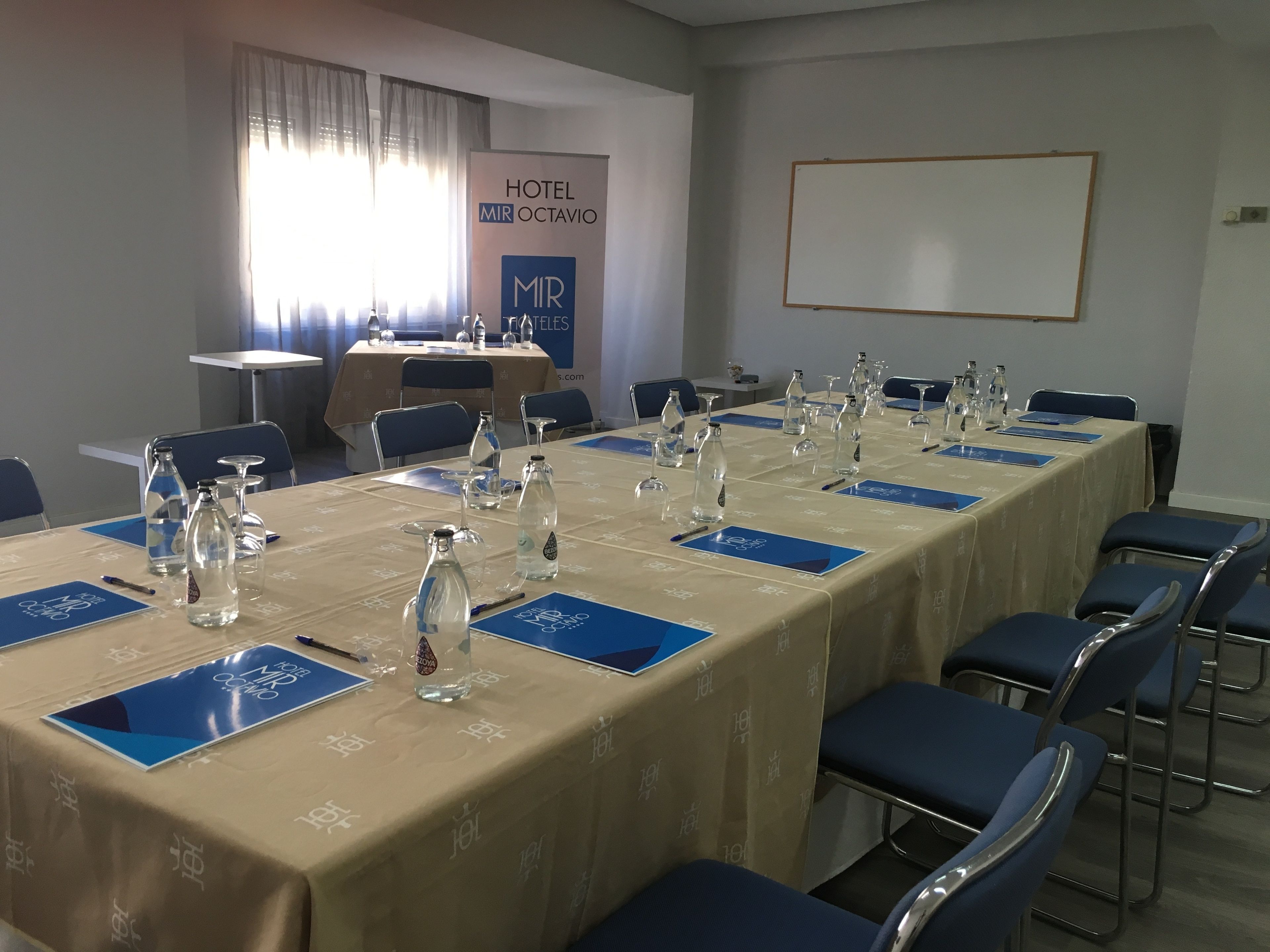 Un salón de conferencias con una mesa larga dispuesta para una reunión, placas con nombres azules y botellas de agua.