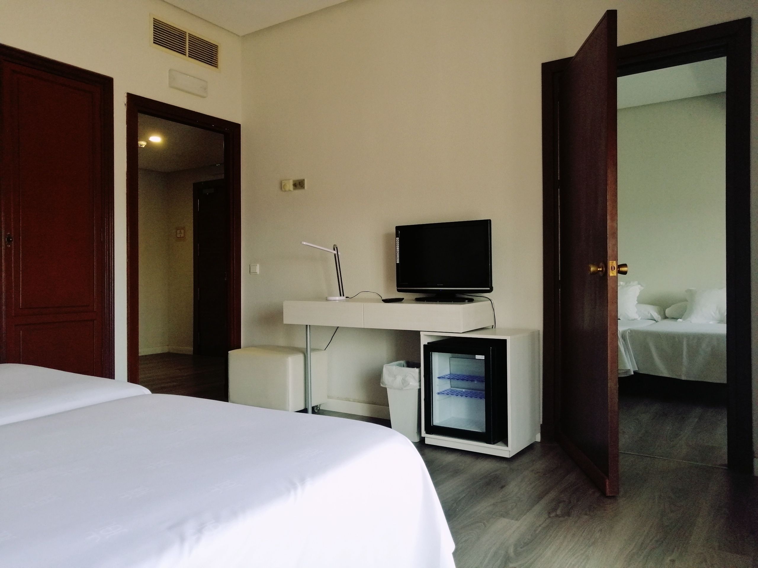 A hotel room with a bed, a desk with a computer and a television, and a small refrigerator.