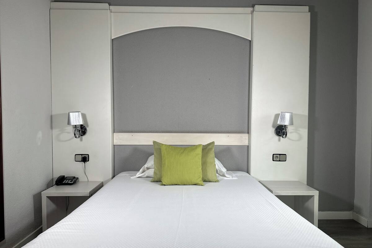 Una cama bien hecha con un colchón blanco y almohadas verdes, flanqueada por dos mesitas de noche con lámparas y un teléfono.