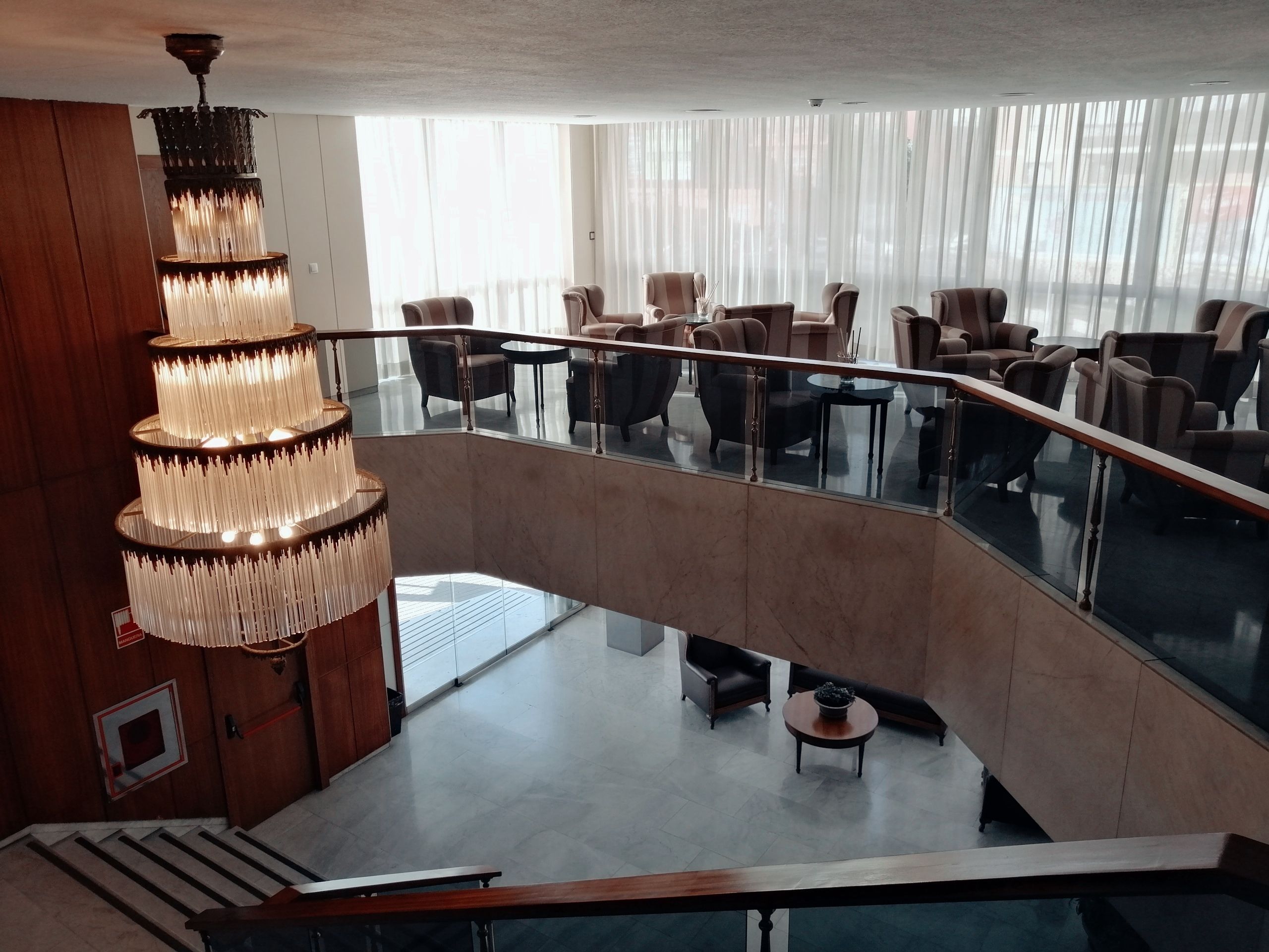Un lobby moderno de hotel con una imponente lámpara de araña, barandilla de vidrio y un amplio espacio de descanso con múltiples sillas y mesas.