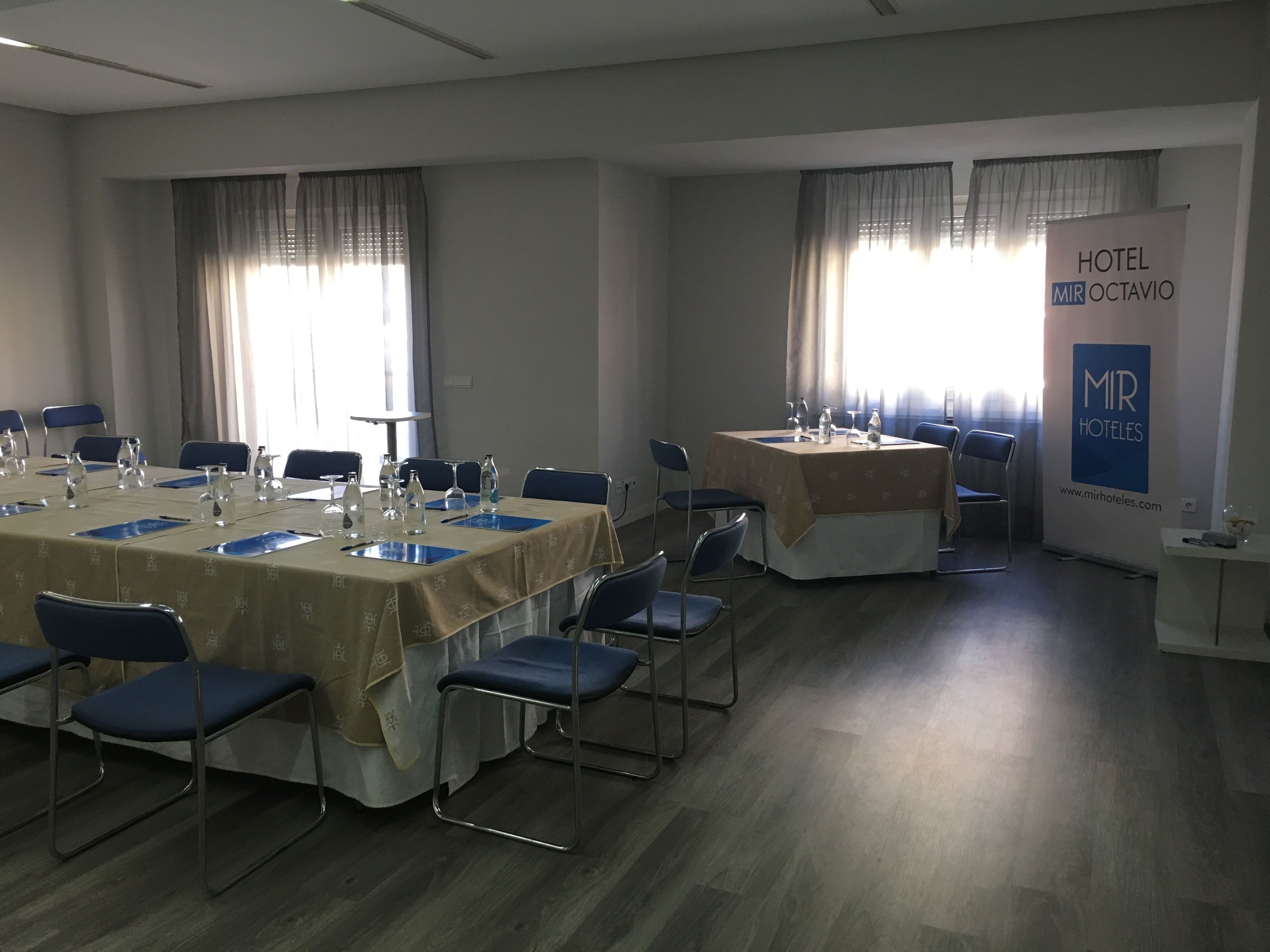Un salón de conferencias con mesas dispuestas para reuniones, equipado con botellas de agua y vasos, y un cartel para Hotel Mir Octavio.