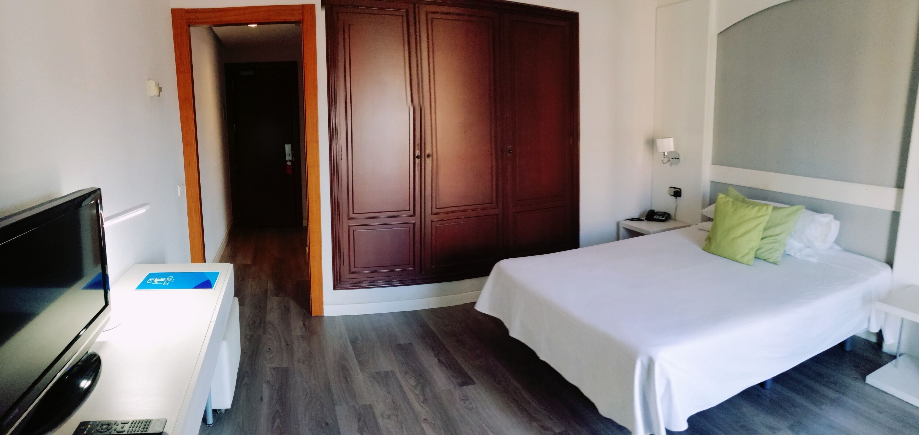 Una habitación de hotel con cama, escritorio con televisor, armario y mesita de noche con lámpara.