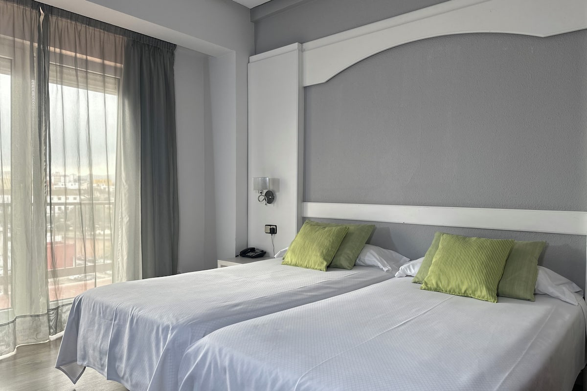 Habitación de hotel con dos camas, una mesita de noche, una lámpara de pared y un ventanal con cortinas translúcidas.