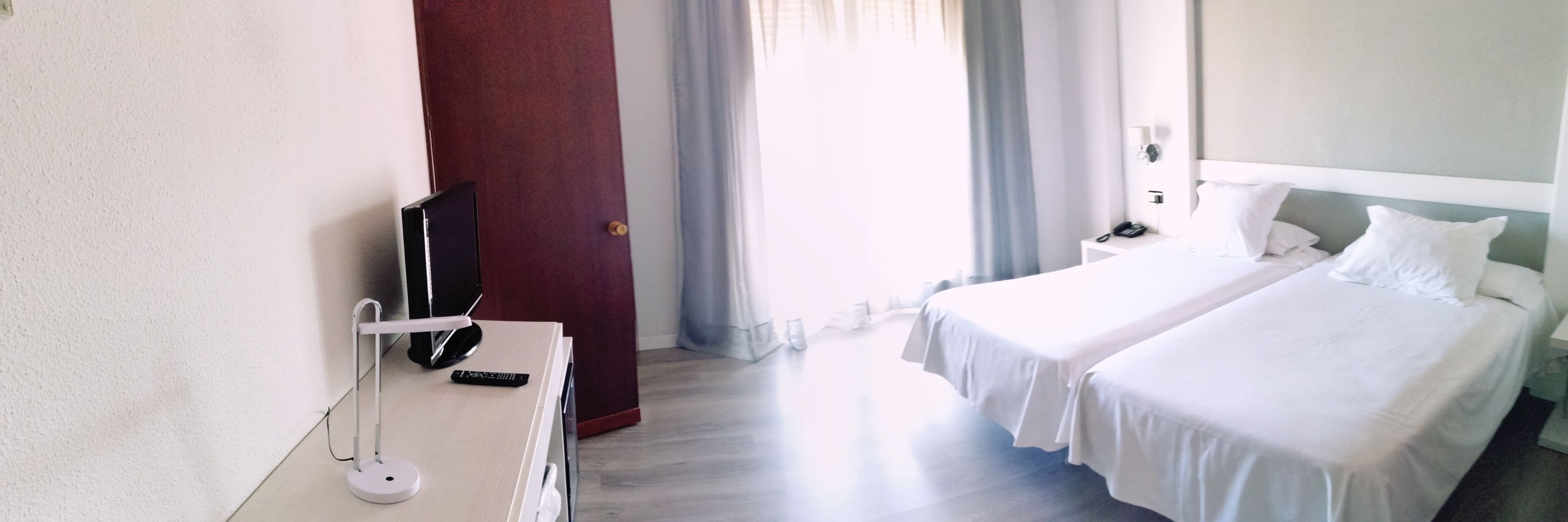 Ett hotellrum med två sängar, ett skrivbord med en lampa, en TV och en dörr som leder ut till en balkong.