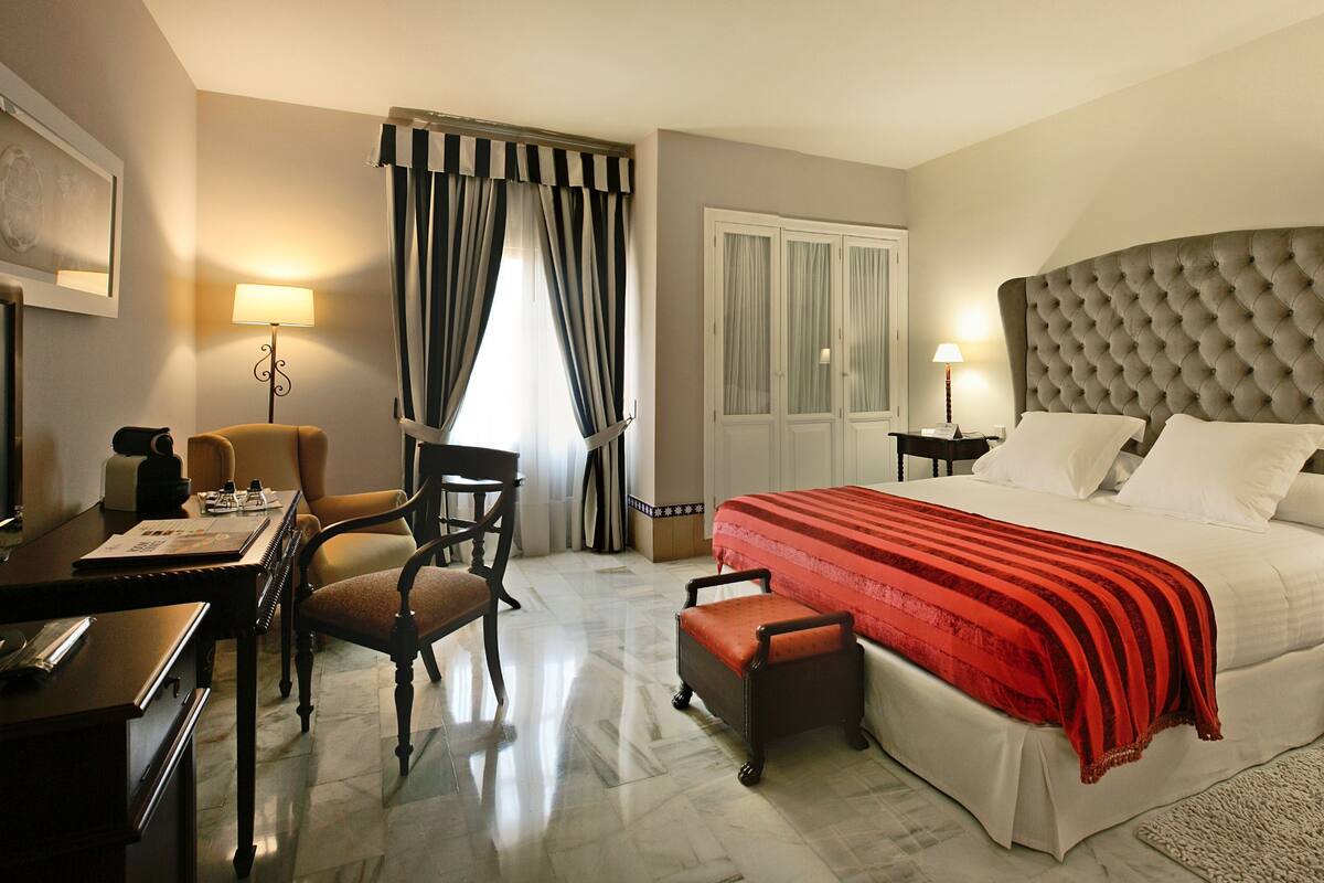 Habitación de hotel con una cama grande, un escritorio con silla, una mesita y una ventana con cortinas.