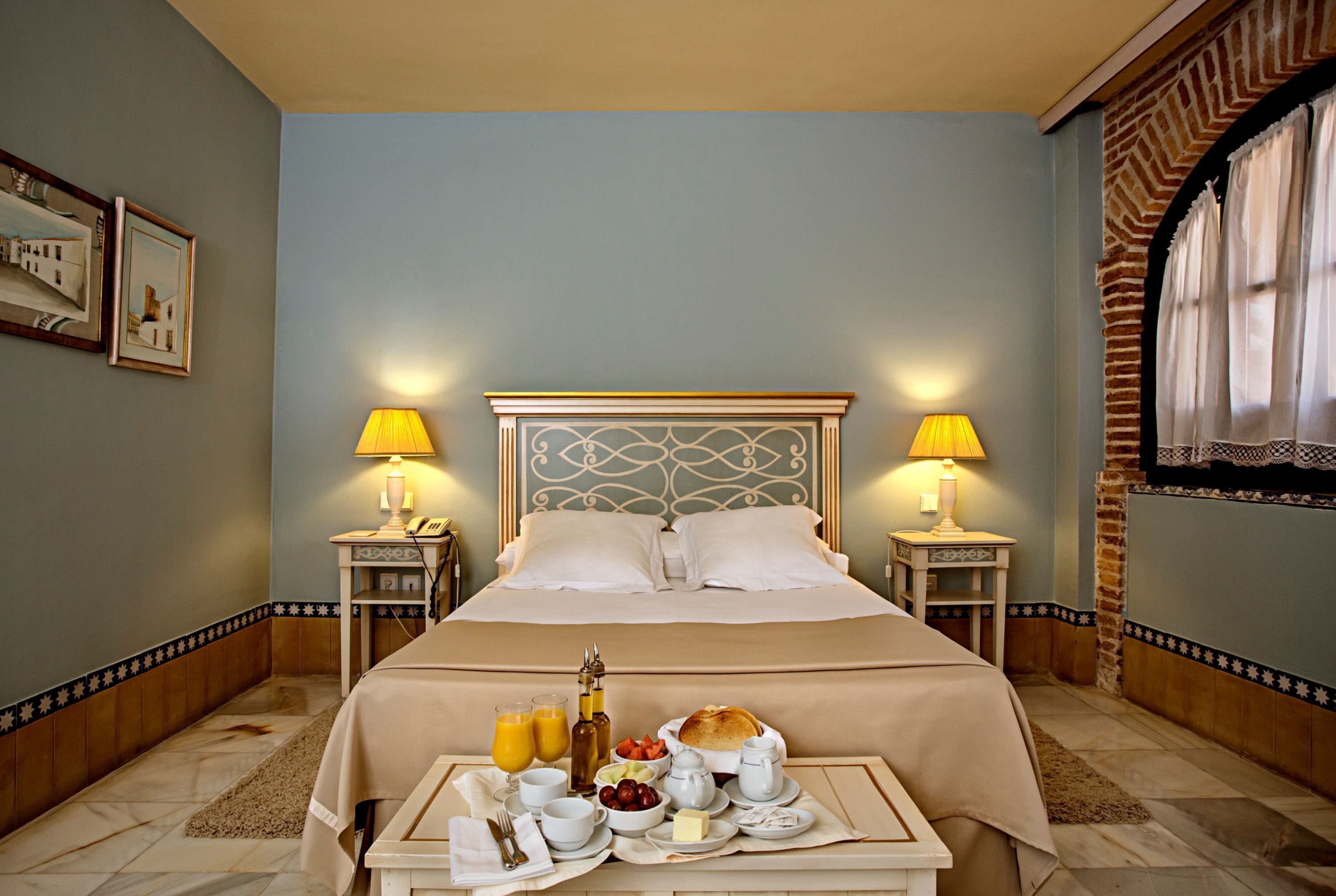 Une chambre d’hôtel avec un lit soigneusement fait, deux tables de chevet avec des lampes, un plateau de petit-déjeuner avec des œufs, des toasts, des fruits et des boissons, et une tête de lit décorative.