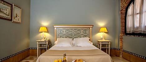 Une chambre d’hôtel avec un lit soigneusement fait, deux tables de chevet avec des lampes, un plateau de petit-déjeuner avec des œufs, des toasts, des fruits et des boissons, et une tête de lit décorative.