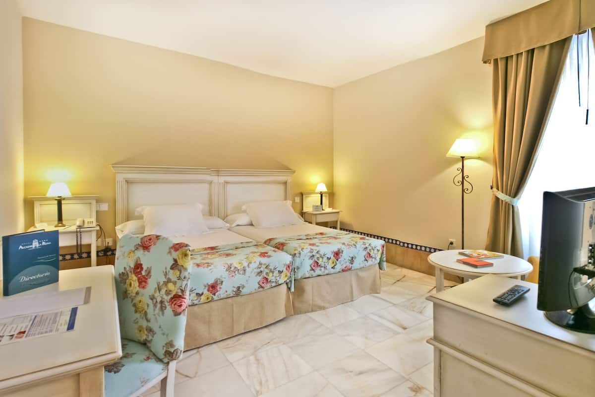 Habitación de hotel con cama, mesitas de noche, escritorio, silla, lámpara, televisor y ventana con cortinas.