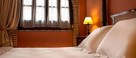 Una habitación de hotel con una cama grande, una ventana con cortinas, una mesita de noche con lámpara y un cabecero de madera.