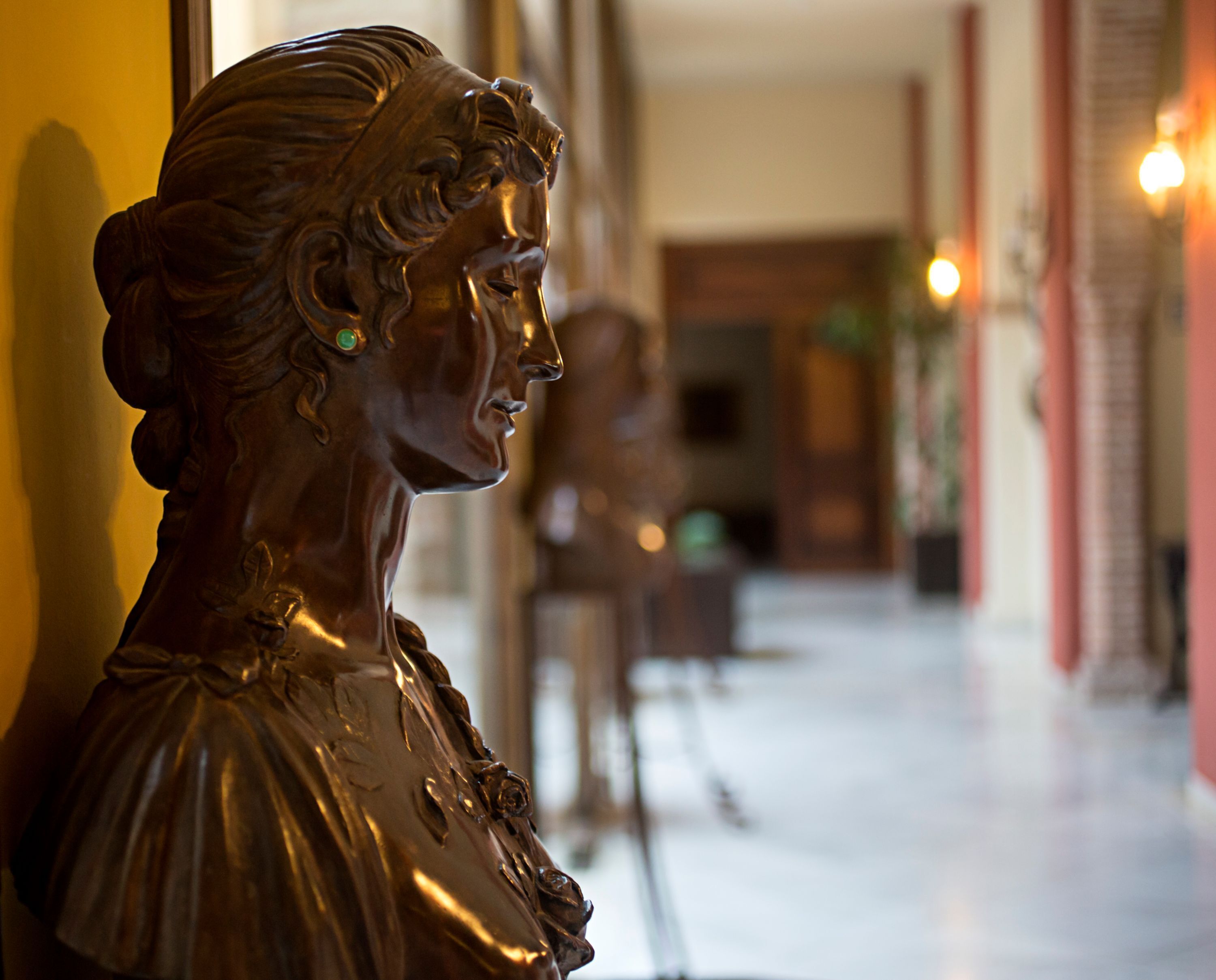 Una escultura de una mujer en perfil, con un fondo borroso que muestra un pasillo y una puerta.