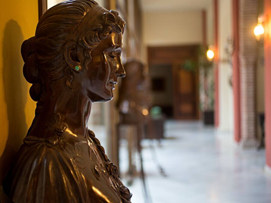 Una escultura de una mujer en perfil, con un fondo borroso que muestra un pasillo y una puerta.