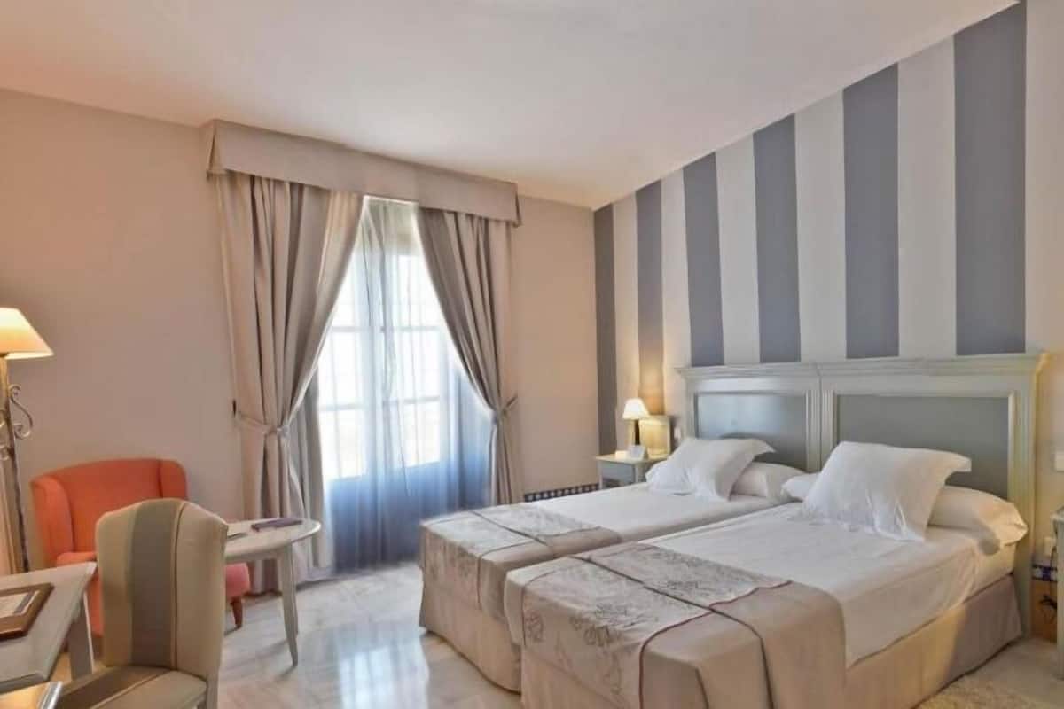Habitación de hotel con dos camas, un escritorio, una silla y un ventanal con cortinas.
