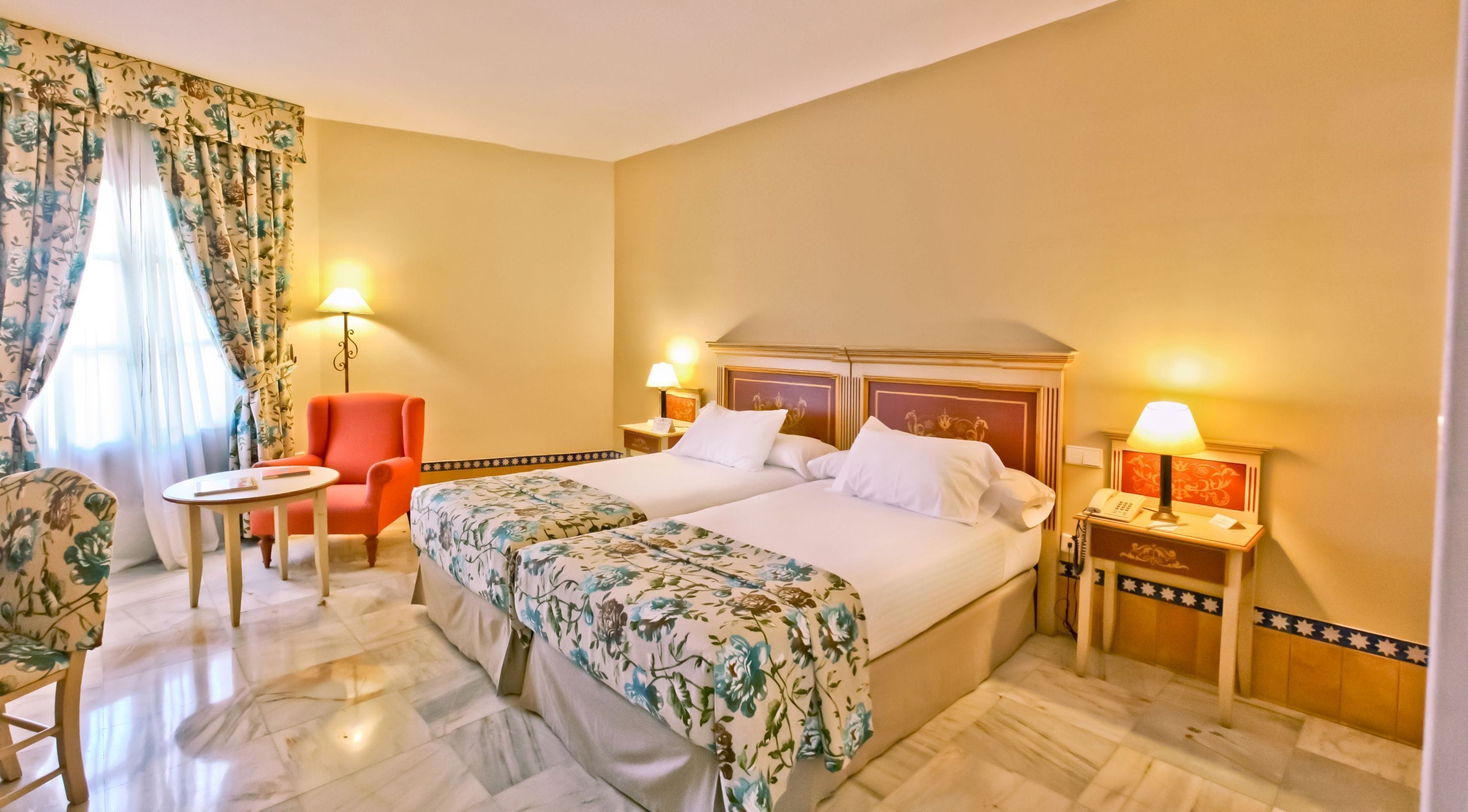 A hotel room with two beds, a chair, a table, and lamps.