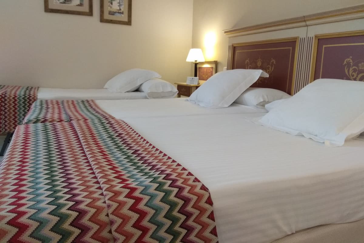 Habitación de hotel con dos camas, una manta estampada, una mesita de noche con lámpara y dos cuadros en la pared.