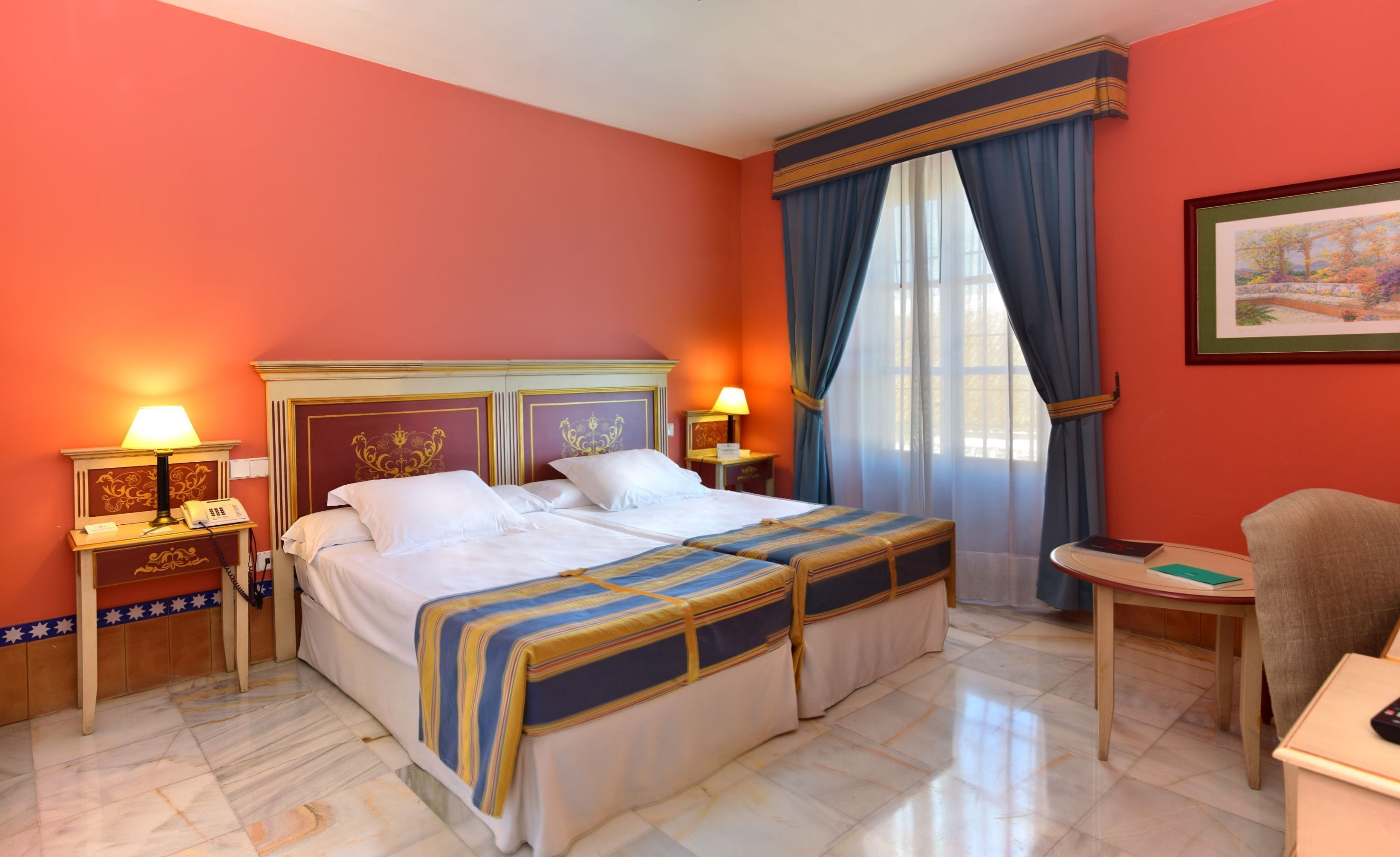 A hotel room with a double bed, two bedside tables with lamps, a chair, a small table, and a painting on the wall.