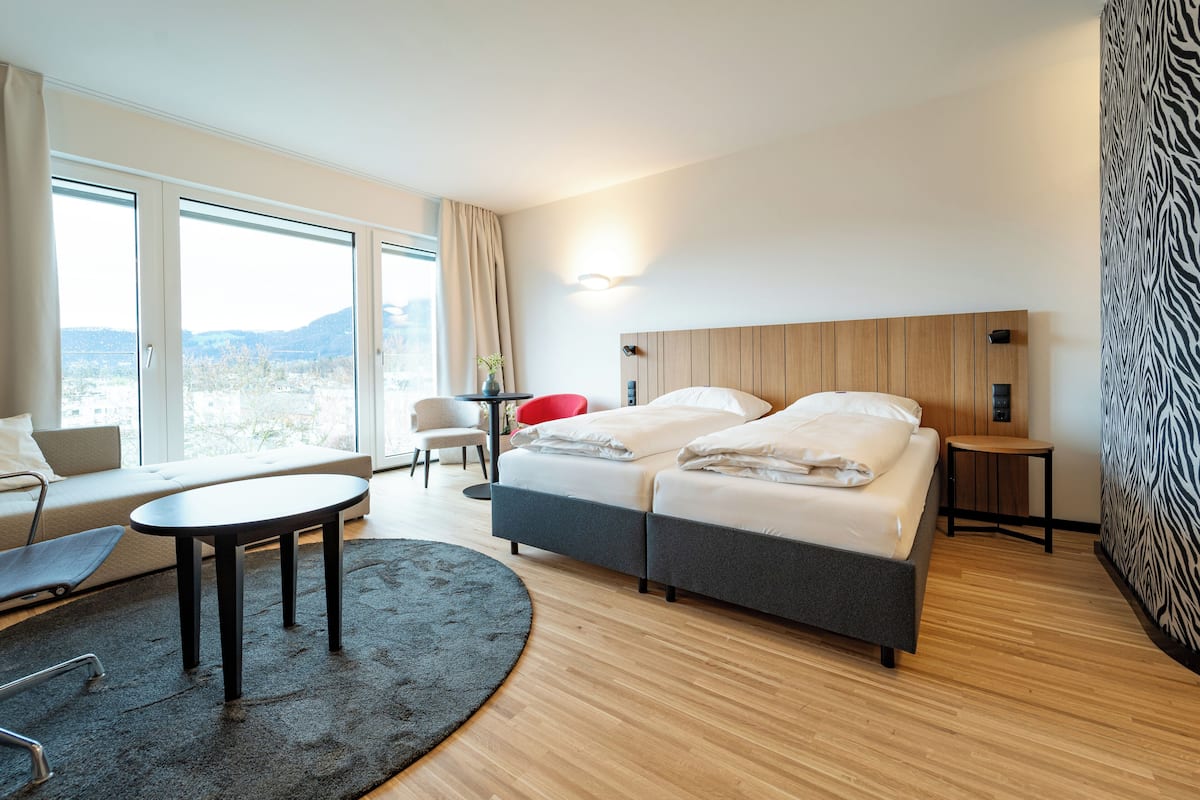 A modern hotel room with a large bed, a sofa, a small table, and a view of the outdoors.