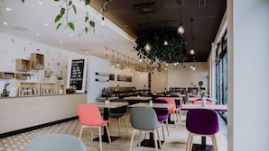 Un café moderno con mesas de madera y sillas coloridas, una planta colgante y una pizarra con el menú.