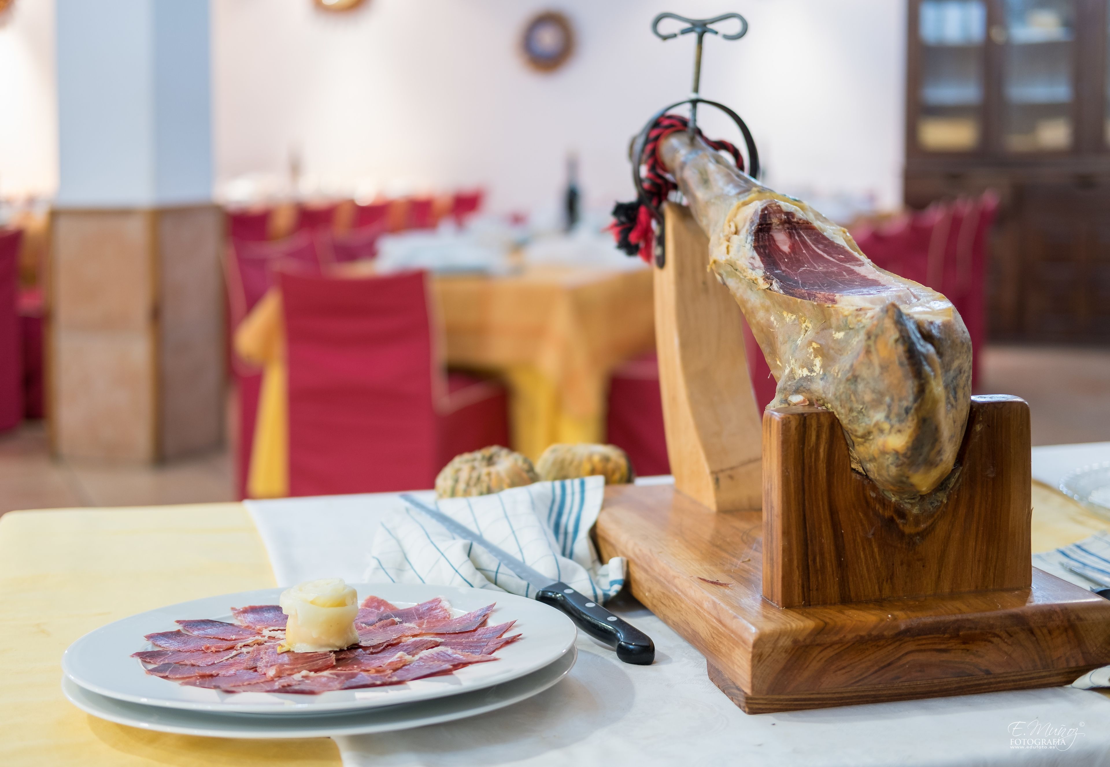 Un soporte de madera para cortar carne con un gran trozo de carne, un plato con carne en rodajas y un pequeño trozo de mantequilla, y un cuchillo sobre una mesa en un comedor.
