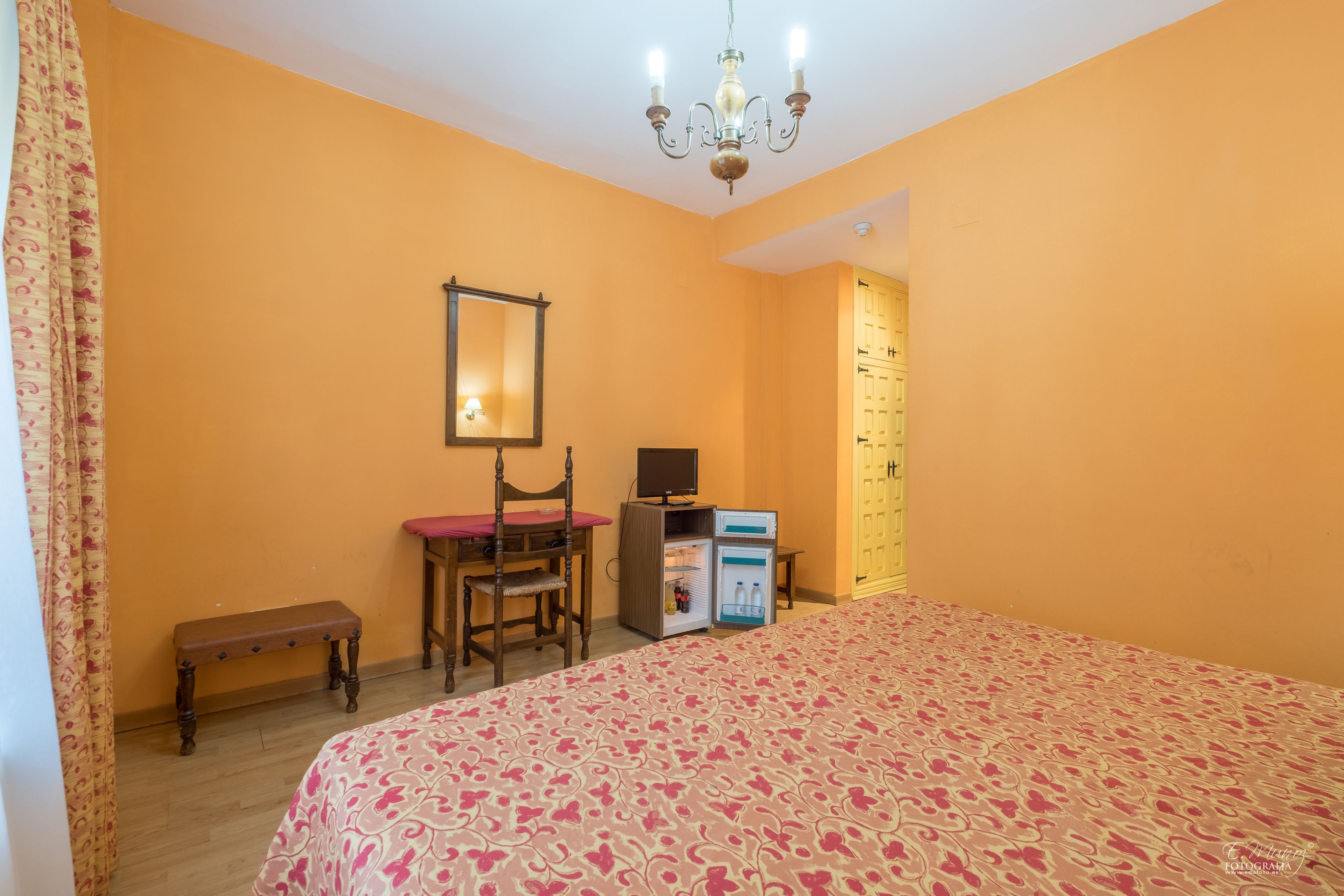 Un dormitorio con cama, escritorio y silla, una pequeña banca, televisión y armario.