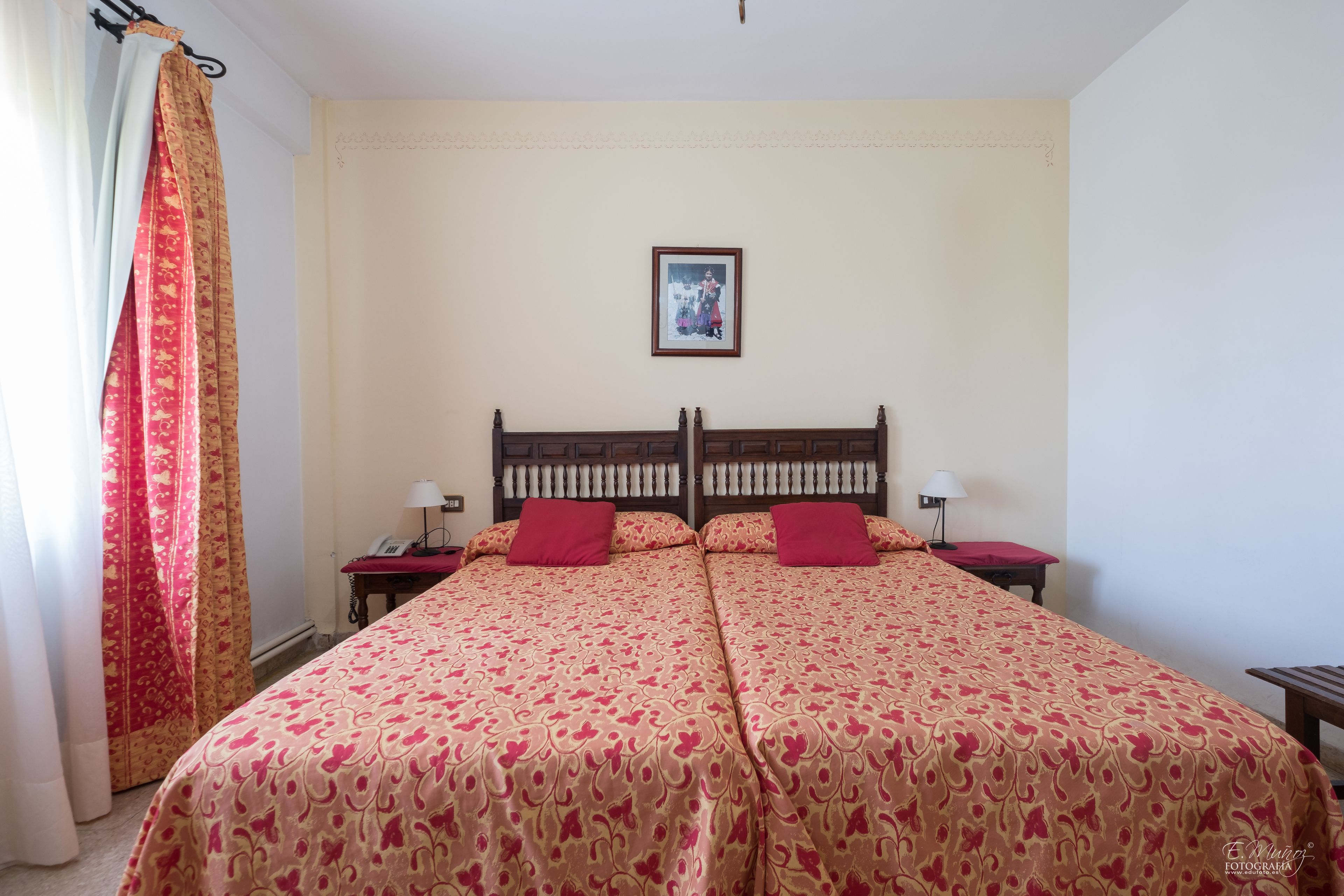 Una cama doble con una colcha estampada, dos mesitas de noche con lámparas, una mesita auxiliar, un cuadro enmarcado en la pared y una ventana con cortinas.